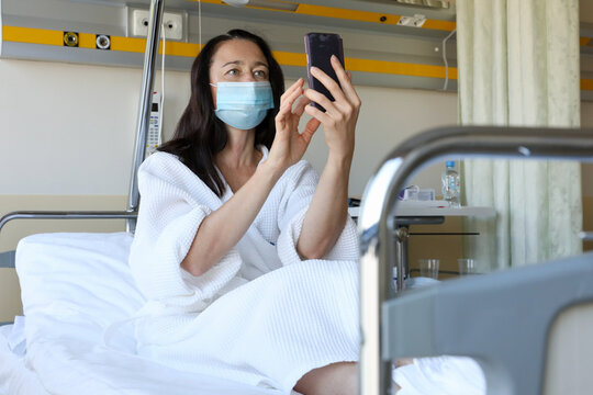 Middle Aged Woman Sitting On Bed In Hospital Ward And Holding Cell Phone In Hand, Chatting, Watching Or Taking Selfie Photo.