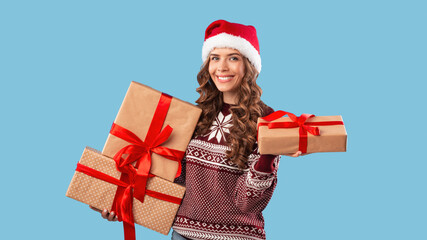 Happy young woman in Santa hat and Xmas sweater holding Christmas gifts on blue studio background, panorama
