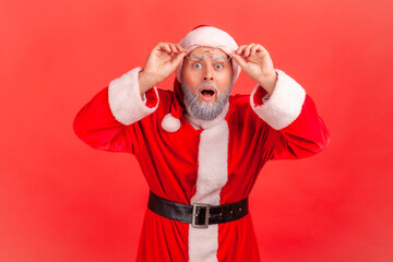 Fototapeta na wymiar Portrait of extremely shocked surprised santa claus looking at camera with big wondered eyes holding eyeglasses on forehead, holidays coming. Indoor studio shot isolated on red background
