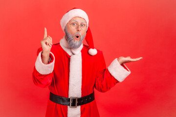 Fototapeta na wymiar Portrait of inspired thoughtful elderly man in santa claus costume pointing finger up, has good idea about holidays celebration, solution. Indoor studio shot isolated on red background