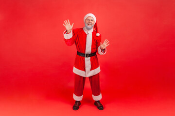 Full length portrait of friendly smiling gray bearded man in santa claus costume raising hands greeting and welcoming looking at camera. Indoor studio shot isolated on red background