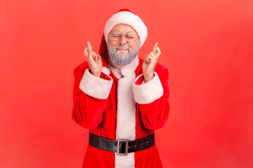 Fototapeta na wymiar Portrait of dreamy elderly man in santa claus costume closing eyes and crossing fingers making a wish, christmas magic, winter holidays. Indoor studio shot isolated on red background
