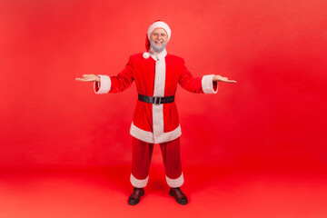 Full length portrait of Kind open hearted elderly man in santa claus costume holding hands opened, giving everything he has, holidays generosity. Indoor studio shot isolated on red background