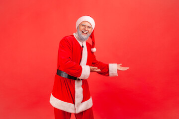 Portrait of positive satisfied gray bearded santa claus showing empty space paying your attention on advertisement area. Indoor studio shot isolated on red background