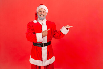 Fototapeta na wymiar Portrait of cheerful santa claus in red costume pointing finger away showing freespace for advertising, looking at camera with toothy smile. Indoor studio shot isolated on red background