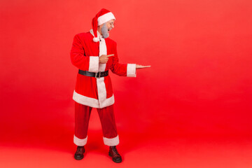Full length portrait of positive happy man in santa claus clothes pointing finger away showing empty space for advertisement, freespace. Indoor studio shot isolated on red background
