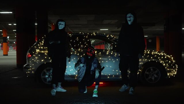 People with luminous purge masks are holding a baseball bat and katana. Killer at night in the parking lot against the backdrop of a car with a garland.
