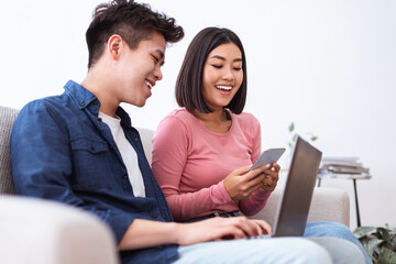 Asian Couple Using Laptop And Smartphone Sitting On Couch Indoors