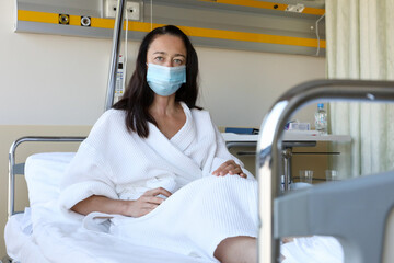 Fototapeta na wymiar Middle aged woman wearing medical face mask and white robe sitting alone on bed in hospital ward 