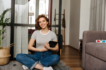 Obraz premium Charming woman with curly short hair holding computer tablet and sitting on floor in living room