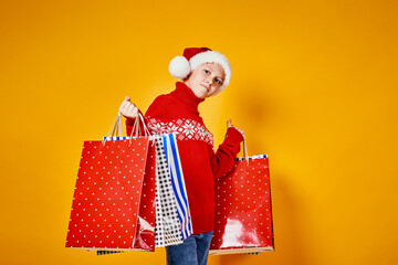 Cute boy in red sweater holding shopping bags with Christmas gifts and looking at camera on yellow...