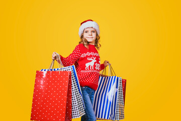 Adorable child in red Santa hat and sweater with deer carrying colorful shopping bags with Christmas gifts on yellow background.