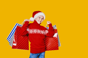 Cute boy in red sweater holding shopping bags with Christmas gifts and looking at camera on yellow background