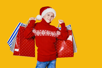 Cute boy in red sweater holding shopping bags with Christmas gifts and looking at camera on yellow background