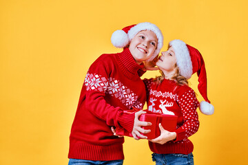 Little girl in red sweater with deer whispering secret to cheerful boy holding gift box on Christmas against yellow background