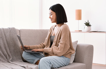 Fototapeta premium Asian woman sitting on couch and working on laptop
