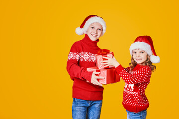 Cheerful kids in Santa hats and red sweaters standing with presents laughing and looking at camera on yellow background