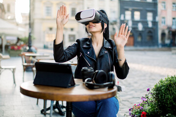 Digital technology, virtual reality. Happy positive young brunette woman, moving hands while trying VR glasses headset during the work or study in the city cafe outdoors