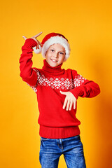 Boy in Santa hat and red sweater having fun on Christmas day against yellow background.