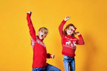 Children in warm red Christmas sweaters and decorated glasses looking at camera on yellow background.