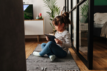 Portrait of girl in jeans sitting on carpet in bedroom and playing in tablet