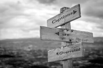 captions are overated text quote on wooden signpost outdoors in black and white.