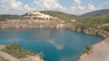 A dazzling lake in the quarry. The white clouds are reflected in the emerald water. Green wooded hills on the horizon.