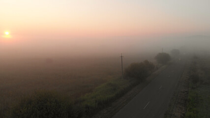 Foggy road near the field. Asphalt driveway covered by dense mist. Cars are riding in white haze.