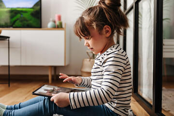 Adorable little girl in good mood watches cartoons in tablet and poses in living room