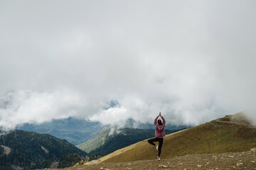 Asana high up in cloudy mountains. Young woman doing yoga stance. Long shot