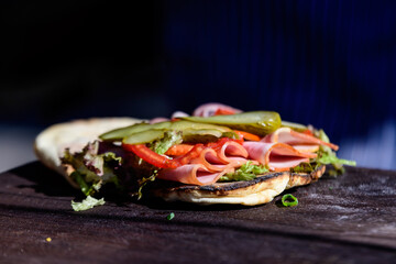 One fresh healthy sandwich or burger with ham, green salad and pickled cucumbers on a brown wooden table in direct sunlight at a street food market, side view photograph with soft focus .