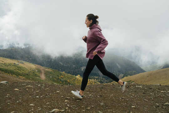 Determined Young Woman Running High Up In Cloudy Mountains. Side View.