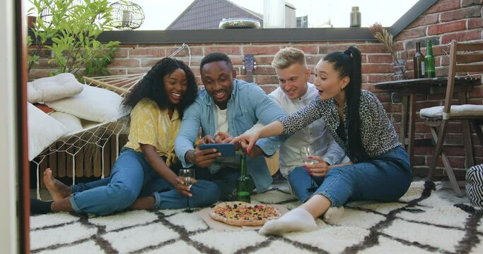 Close up of likable satisfied carefree multiracial group of people which resting on the balcony and browsing funny photos on smartphone during summer party