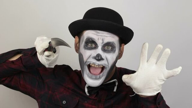 A horrible man in clown makeup threatens his victim with a sharp knife. The scary clown looks at the camera and laughs terribly and threatens his victim with a knife. Halloween costume.