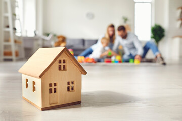 Little wooden house on floor of cozy room with happy family playing in background - obrazy, fototapety, plakaty