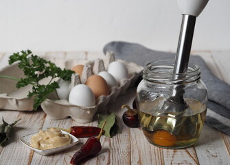 The stage of making homemade mayonnaise from natural healthy ingredients.A jar with ready made mayonnaise, vegetable oil, eggs and a blendar for whipping on a wooden white background.