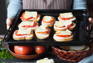 Elderly woman cooks hot sandwiches with cheese, tomatoes, sausage on toast and keeps them on a black baking sheet from the home oven. Healthy natural food.