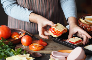 An elderly woman makes hot sandwiches with cheese, tomatoes, sausage on toast. She puts the sandwiches on a baking sheet for the kitchen oven. Healthy natural food.