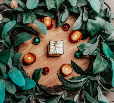 A Flatlay Of A Wreath Framing A Single Gold Wrapped Present And Candles And Ornaments.