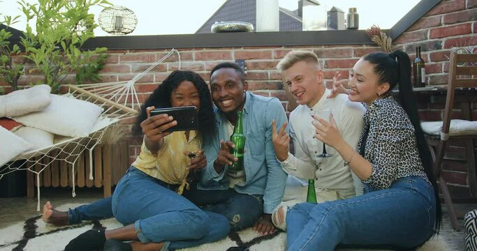 Likable happy cheerful multiethnic youth having fun together while have video call on phone with joint friend during friends meeting on the terrace