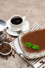 Traditional Italian Tiramisu dessert in glass baking dish, cup of espresso and mocha coffee maker on concrete background