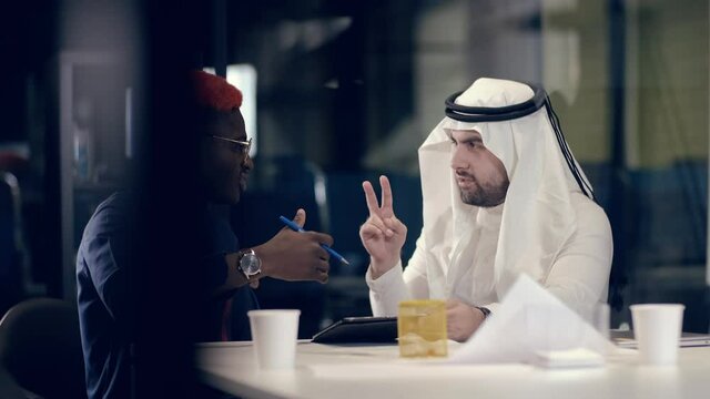 Arab businessman in kandur and his African colleague in national dress have a business conversation while sitting at a table in a modern meeting room. Team cooperation concept. Colleagues work