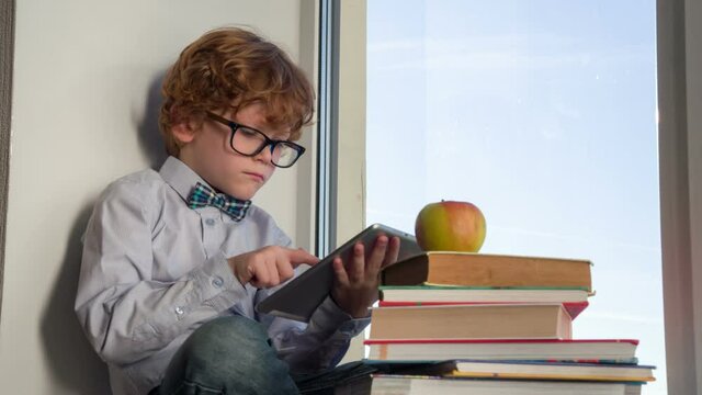 The Little Scientist. Auburn curly boy with glasses sitting on a windowsill with a tablet and books. In the pile of books lies the big apple. The boy looks at him thoughtfully, bites off a piece