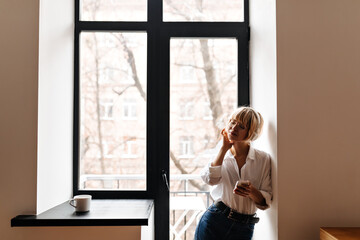 Fototapeta na wymiar Girl in jeans and white shirt listening music in headphones. Indoor shot of blonde european woman with smartphone posing at home.