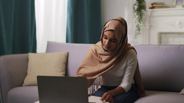 Muslim young woman in hijab scarf and headset is studying online, taking notes. Indian lady freelancer working on computer from home, browsing the web, writing.