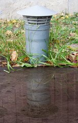 Gray ventilation pipe against a background of green grass and yellow leaves and its reflection in the water