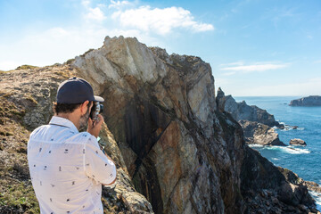 Fototapeta premium Male tourist taking pictures of the ocean on top of a high cliff. He is using a retro style photography camera. Summer time. Cape Penas (Cabo de Peñas) is the most northerly point of Spain, Asturias