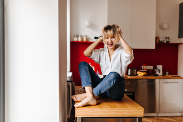 Excited barefoot girl sitting on table. Blissful blonde woman touching hair while posing at home.