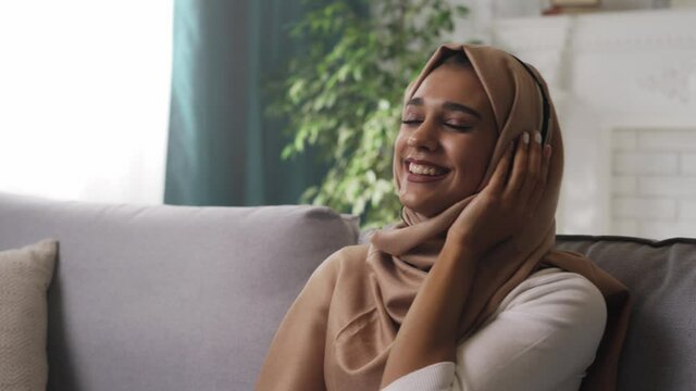 Young modern Indian woman student wearing hijab scarf is enjoying free time at home, relaxing on couch, listening to music in headphones, dreaming with closed eyes. Happy and pleased muslim lady.