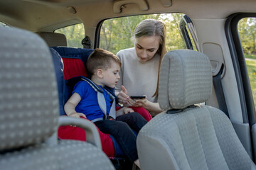 Mother gives the phone little boy sitting in a car seat. Safety of transportation of children.